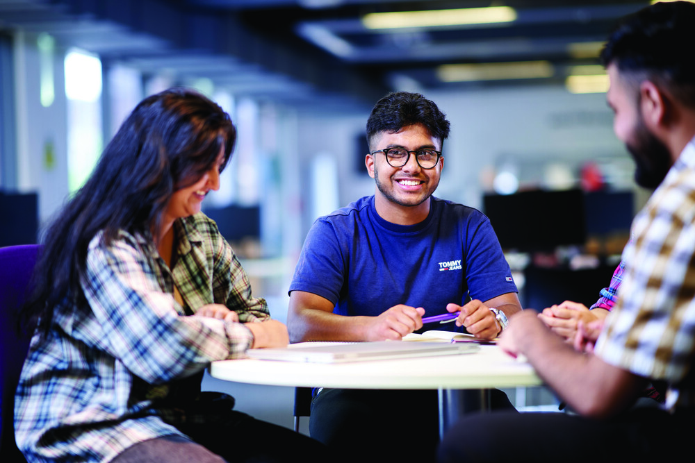 Three students sitting round a small beige round table.  All the three adults are smiling