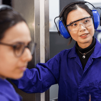 Two engineering students wearing blue lab coats and ear defenders